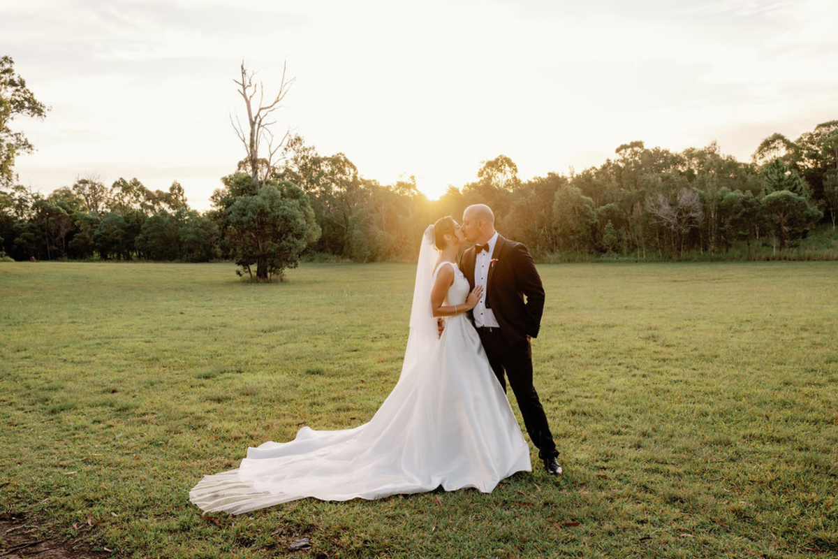 A bride in a long white wedding gown with a veil and a groom in a black suit embrace and kiss on a grassy field at sunset, with trees in the background.