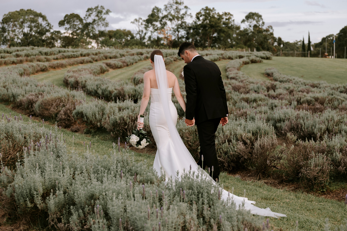 A bride in a white sleeveless wedding gown with a long train and veil walks hand-in-hand with a groom in a black suit through a lavender field.