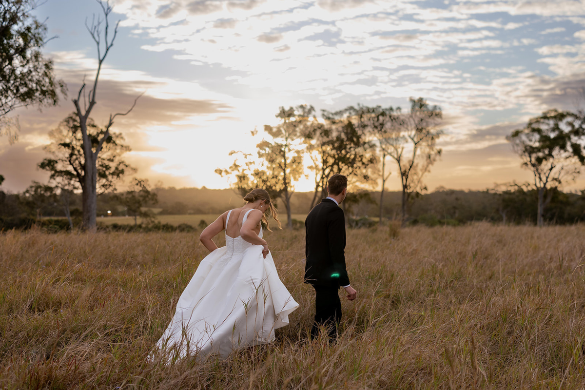 A bride in a white wedding gown and a groom in a black suit walking through a dry grass field at sunset, with trees and a cloudy sky in the background.