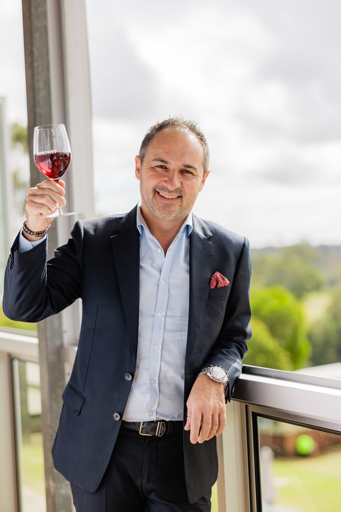 Man in a suit holding a glass of red wine on a balcony with a scenic background