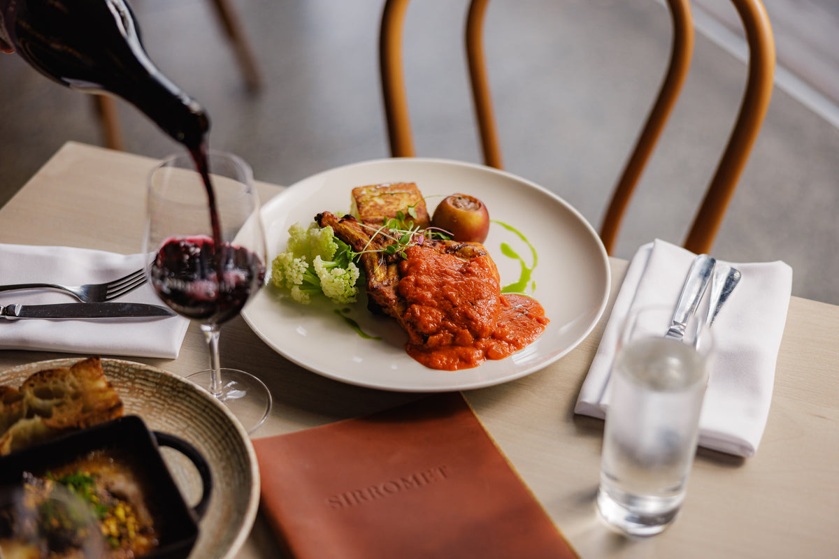 A plated gourmet dish with red sauce, greens, and a small apple on a restaurant table, with a wine bottle pouring red wine into a glass and a water glass nearby.