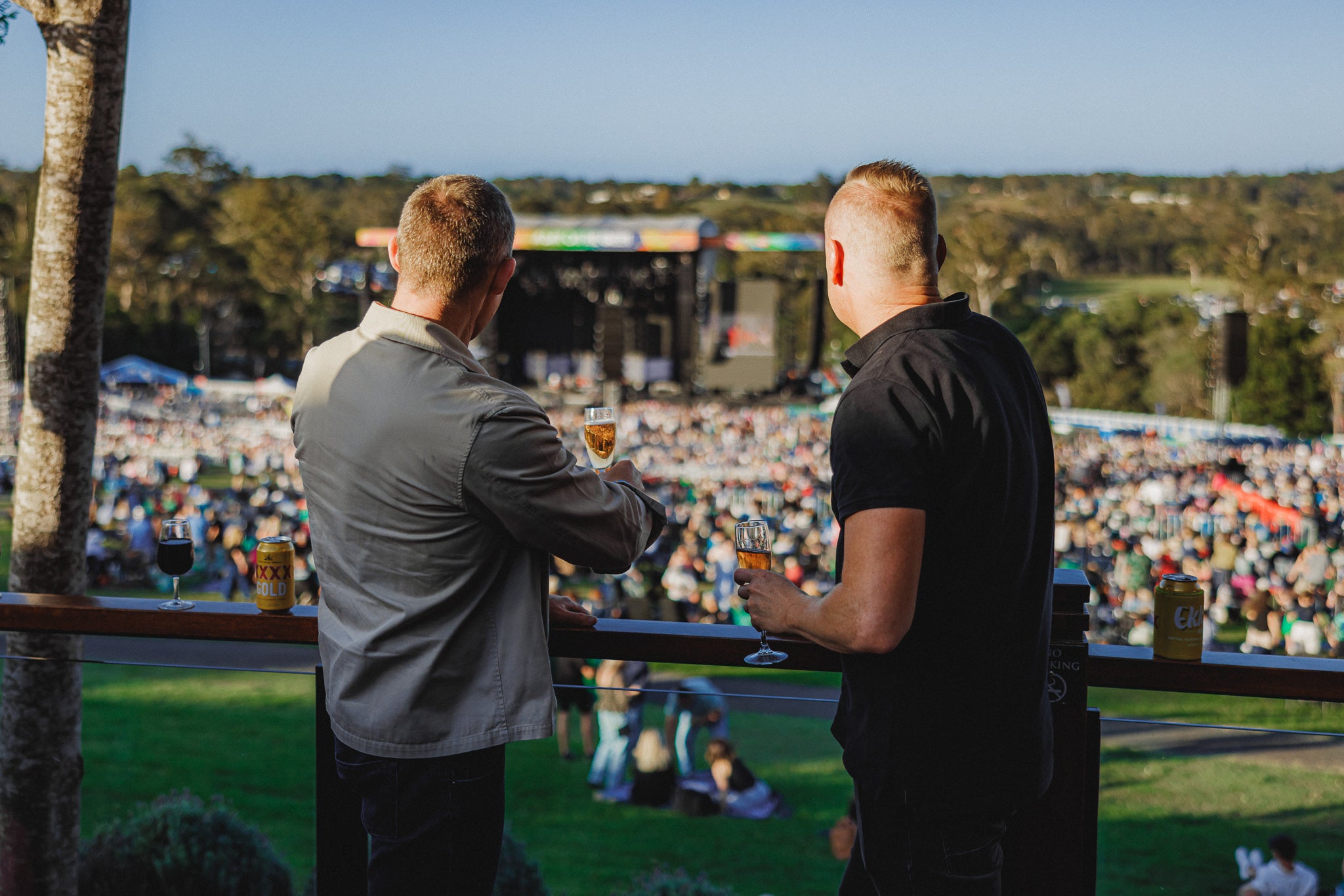 Two men standing at a concert venue, one holding a drink, with a crowd and stage in the background.