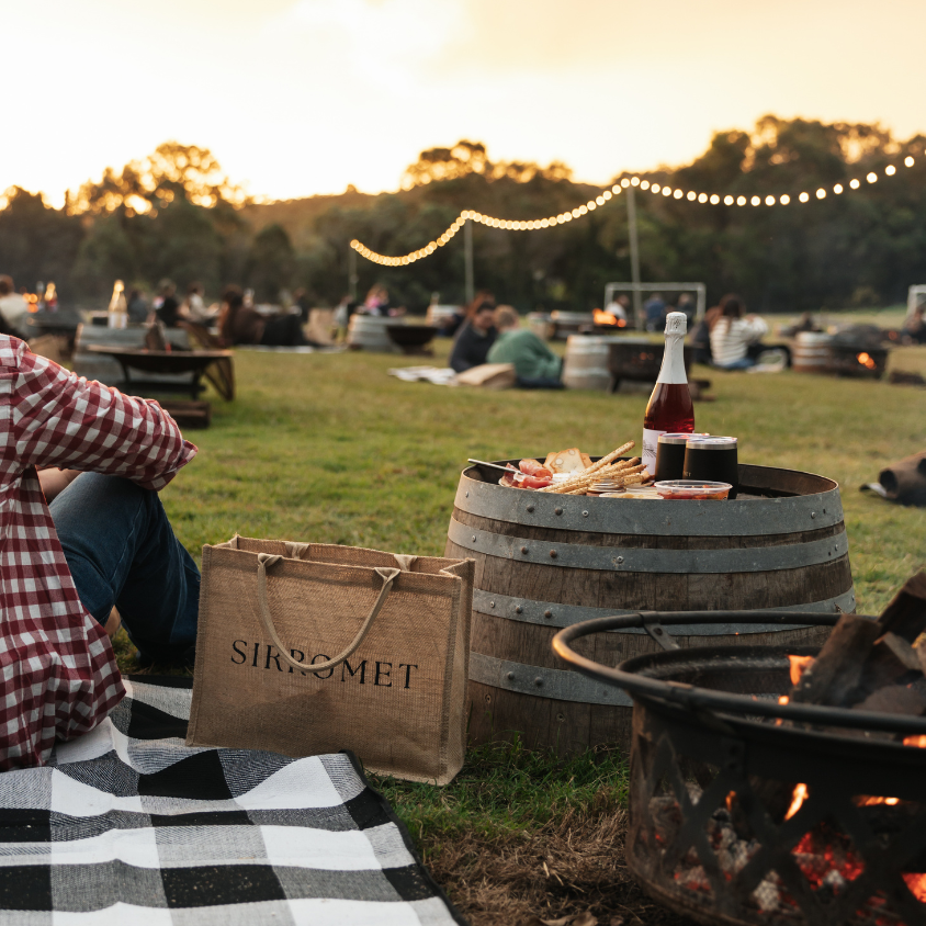 Person sitting on a blanket by a fire pit with a bottle of wine and a bag, surrounded by a casual outdoor setting.