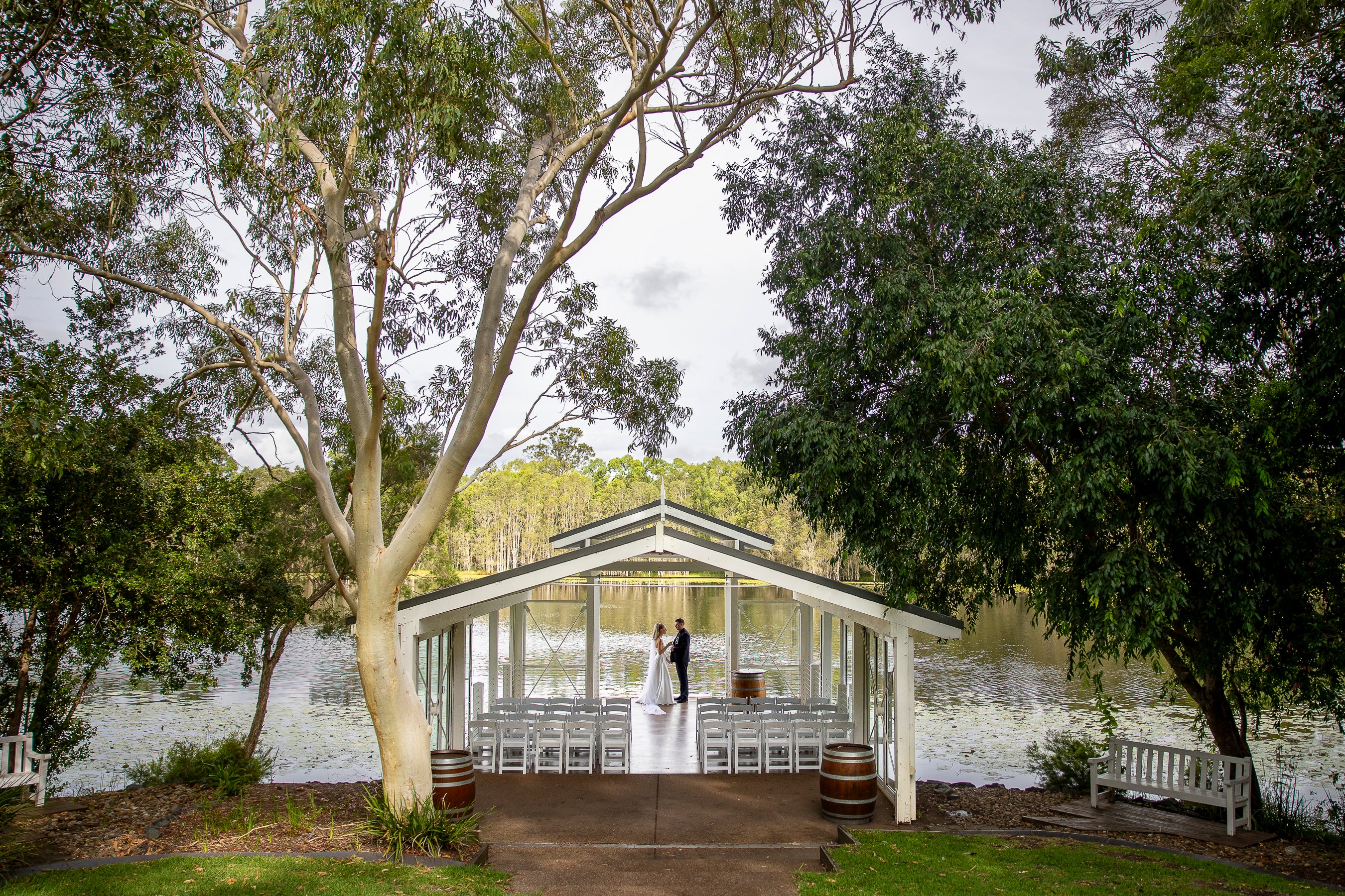 Wedding ceremony taking place in a pavilion by a lake with trees surrounding it.