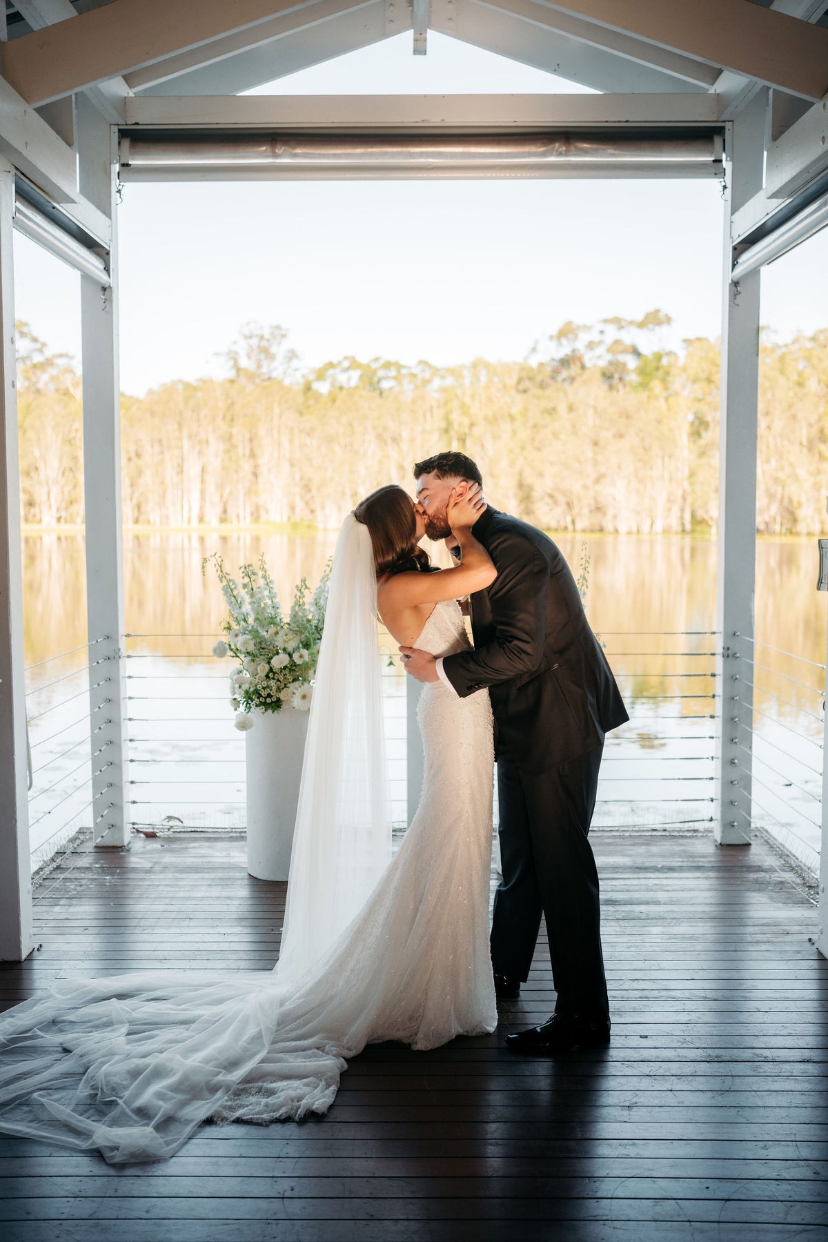 A bride in a white wedding gown and veil and a groom in a dark suit kiss on a wooden dock overlooking a calm lake with trees in the background.
