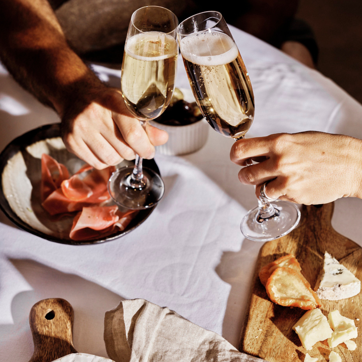 Two hands holding champagne flutes over a white tablecloth, with a plate of sliced cured meat and a cutting board with cheese in a warm, rustic setting.