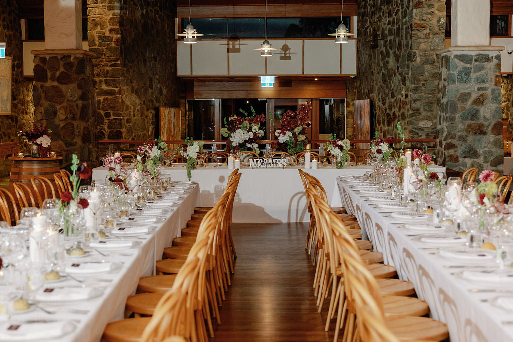 A long banquet table setup in a rustic stone-walled hall, with white tablecloths, wooden chairs, glassware, and floral centerpieces arranged symmetrically down the center aisle.