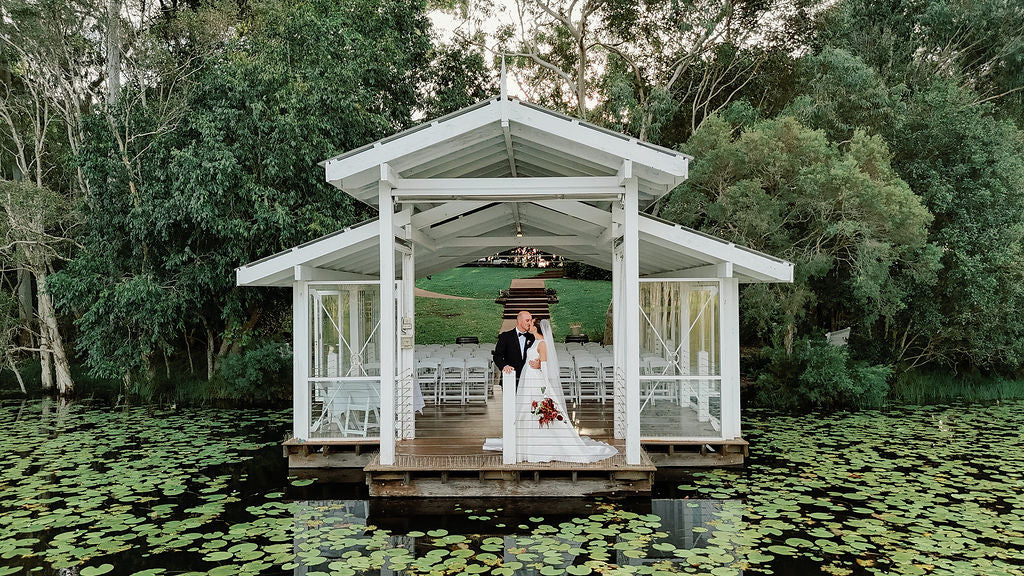 A bride and groom stand under a white wooden dock pavilion surrounded by lily pads on a lake, with a wedding ceremony setup inside and dense green trees in the background.
