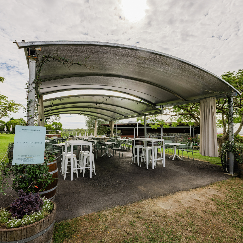 Outdoor event pavilion canopy with white tables and stools arranged underneath on a grassy lawn, under a cloudy sky.
