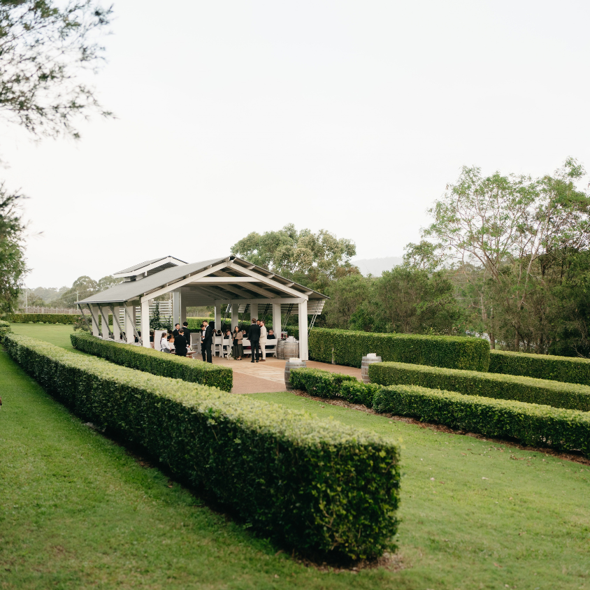 A large outdoor event pavilion with a covered seating area and bar, surrounded by manicured green hedges and grass, with trees in the background under a bright overcast sky.