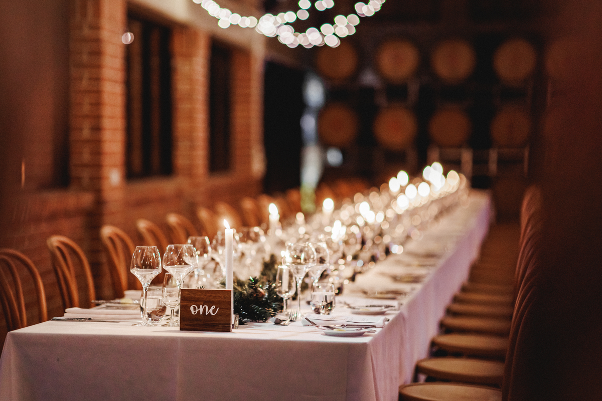 A long wedding reception table set with white tablecloths, clear wine glasses, lit candles, and a small wooden 'one' place card, photographed in a warmly lit event space with brick walls and hanging lights.