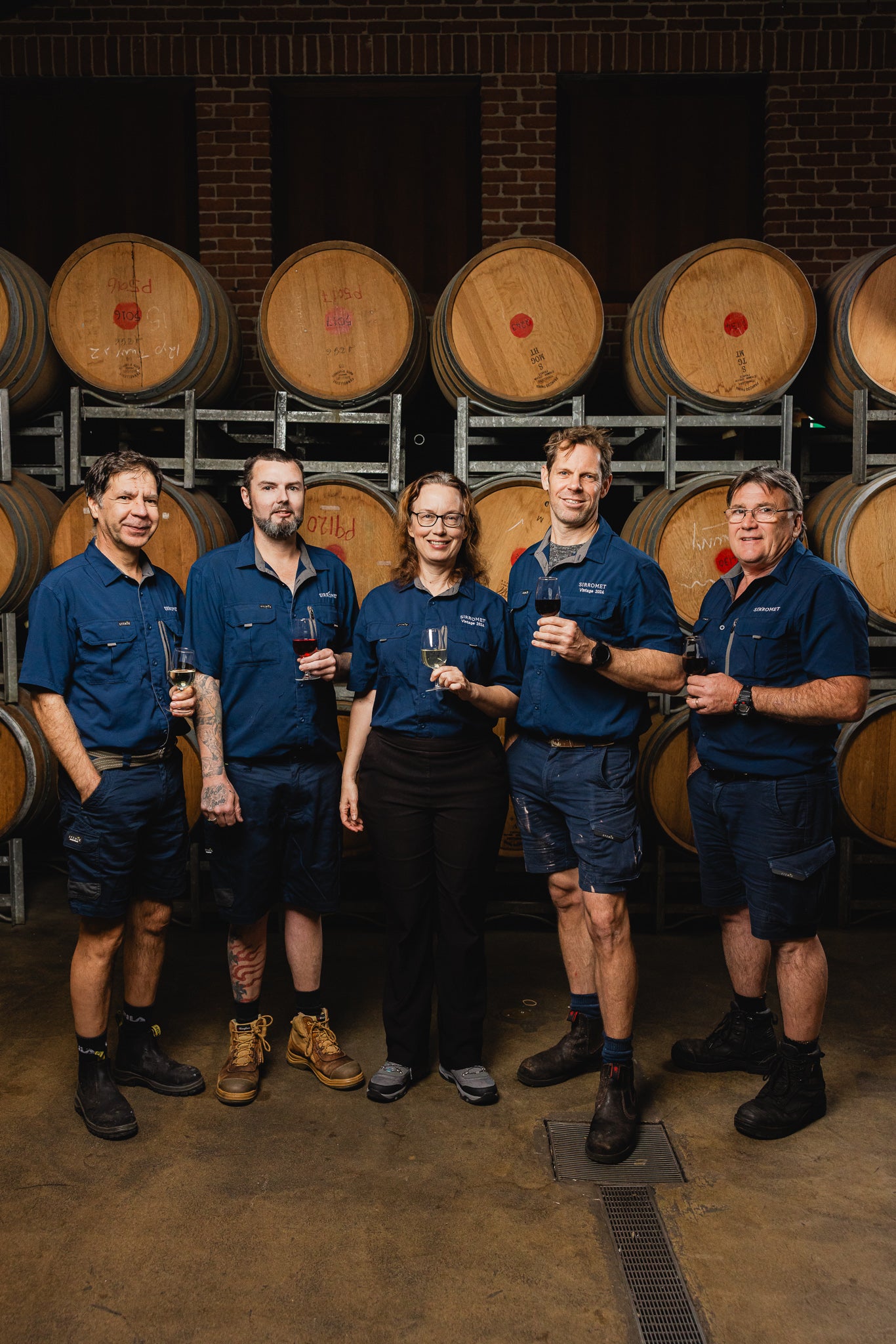 Five people in matching blue uniforms standing in front of wine barrels in a winery.