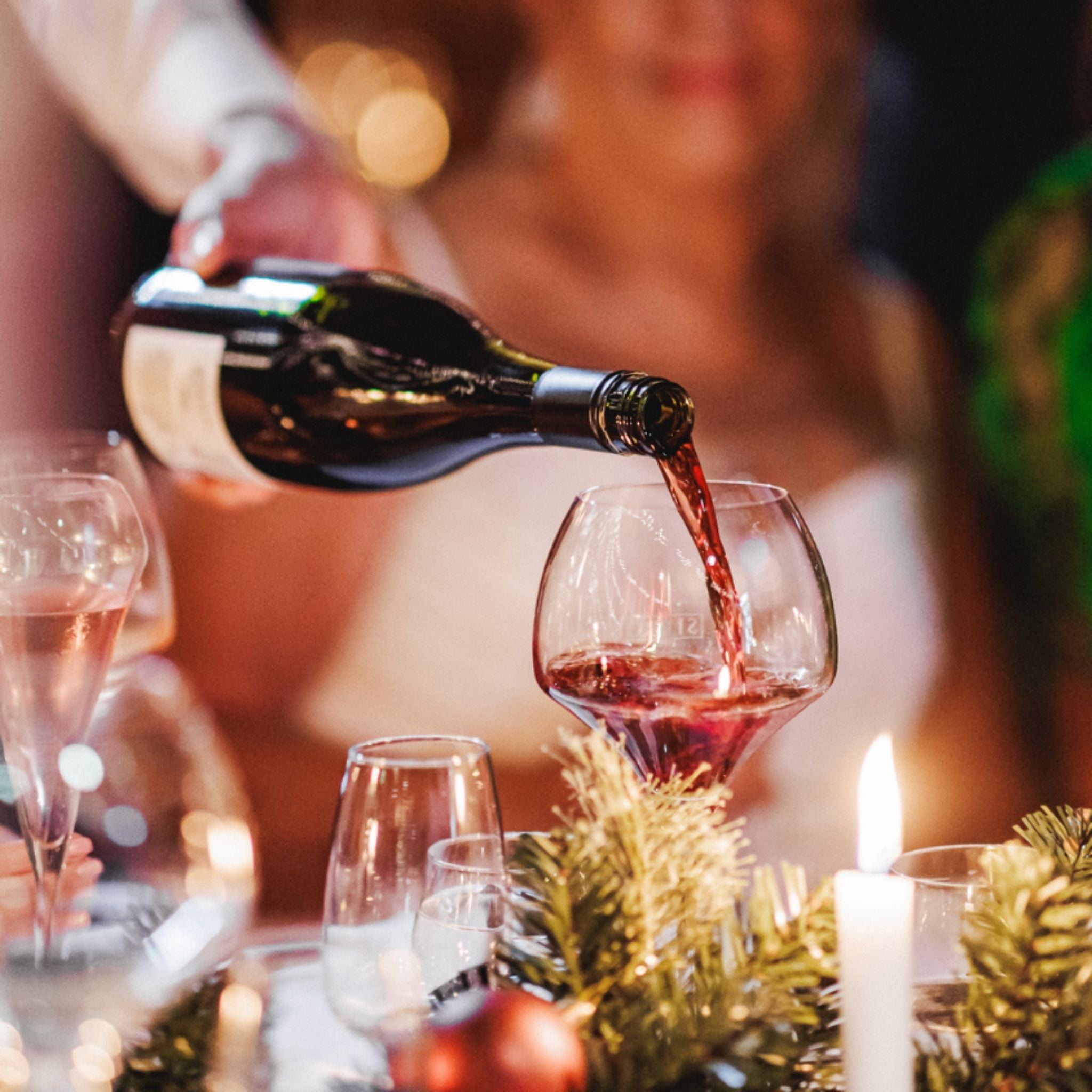 Person pouring red wine into a glass at a festive table with candles and greenery.