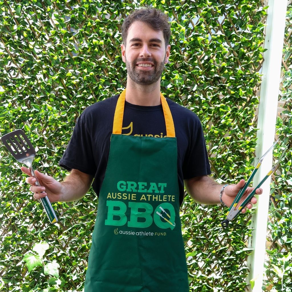 Dominic Bechard wearing a green apron with 'Great Aussie Athlete BBQ' text, holding barbecue tools against a leafy green background.