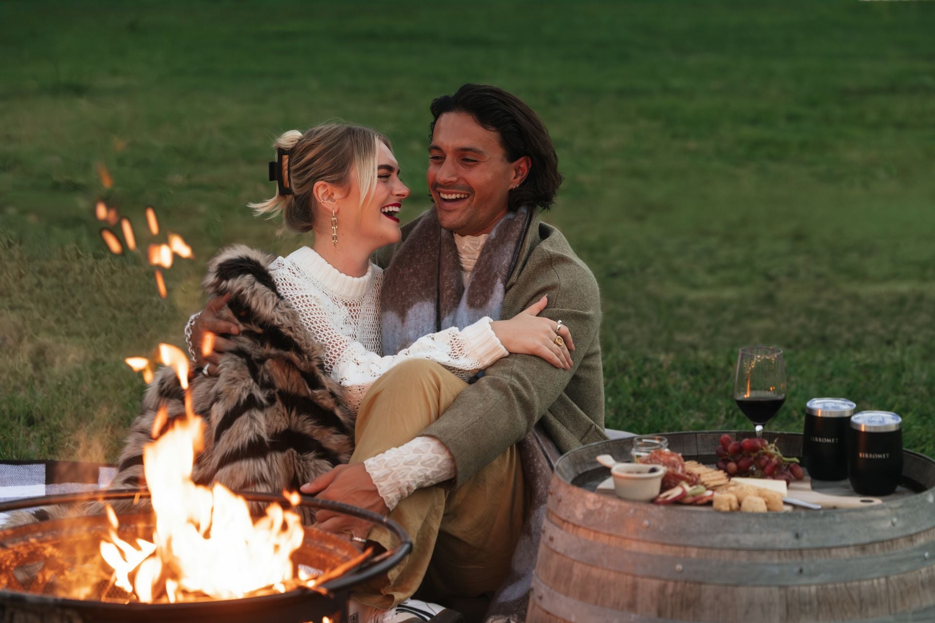 A couple cuddling in front of a fire pit next to wine and cheese platter