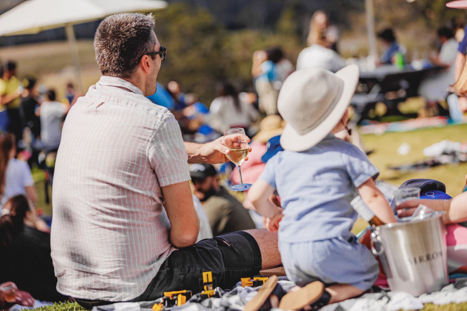 Man and child sitting on a blanket at an outdoor event, enjoying wine.