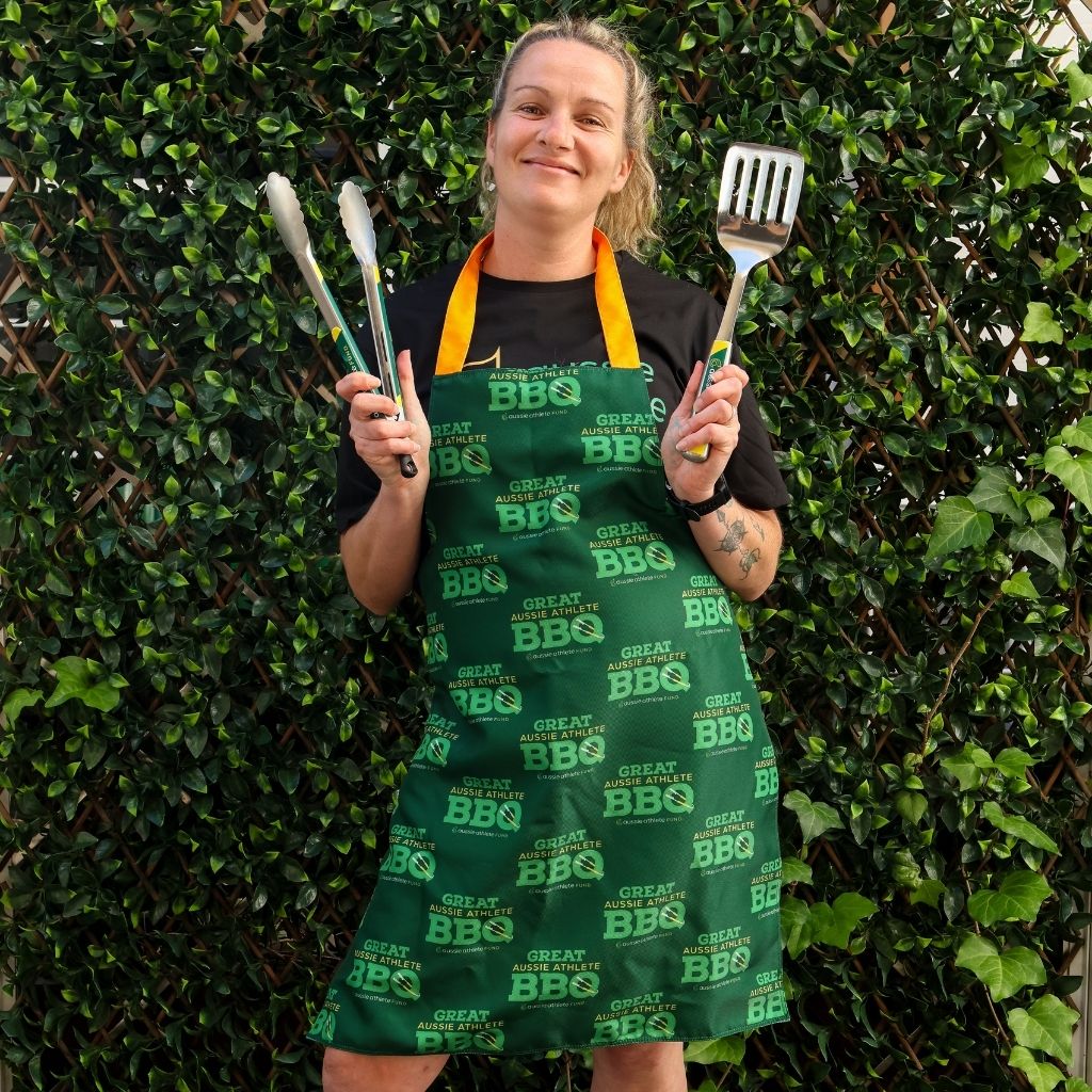 Justeen Kruger wearing a green apron with BBQ text, holding utensils against a green hedge background