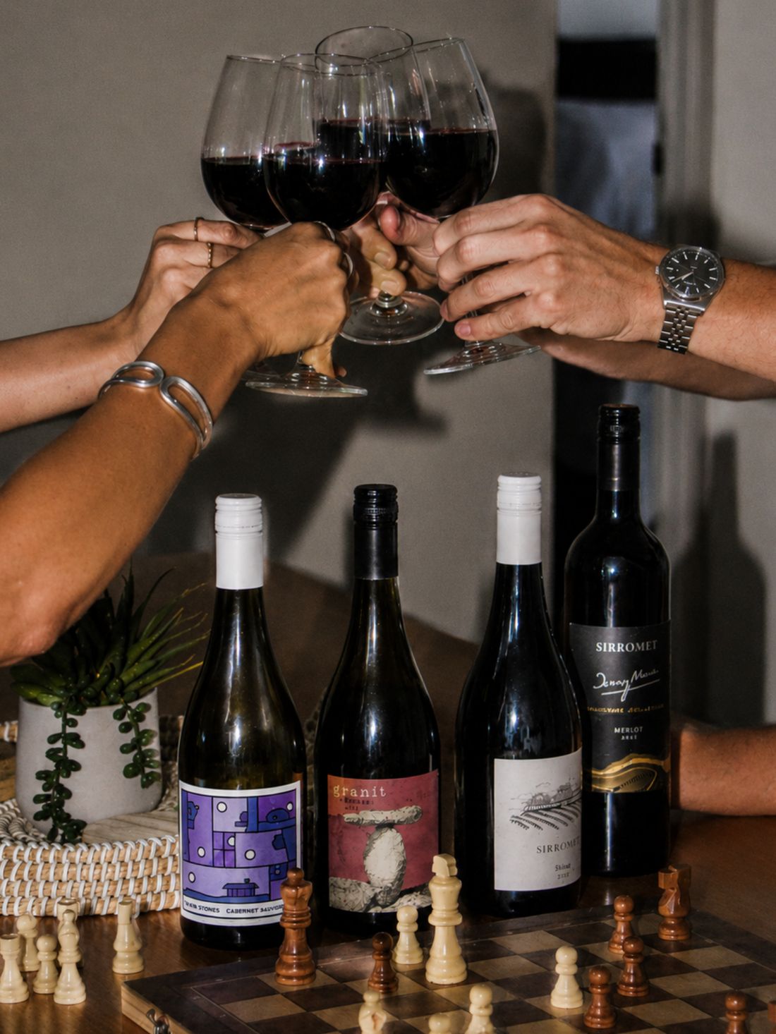 Four wine bottles arranged on a table with a chessboard and playing pieces, while three people's hands and arms raise wine glasses filled with red wine in a toast. The scene is indoors with warm lighting and a neutral wall background.