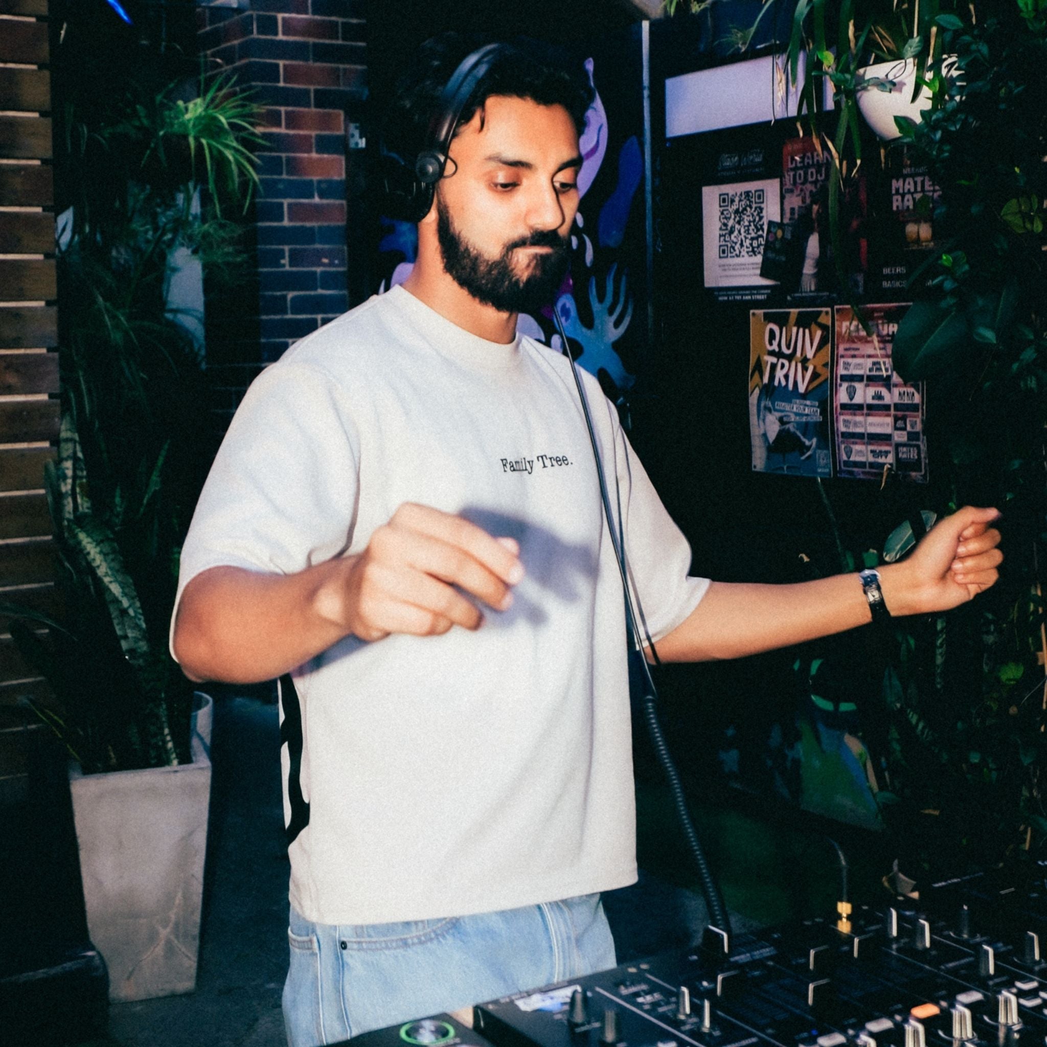 Man wearing headphones and a white t-shirt with text, standing behind DJ equipment in a dark room with plants.